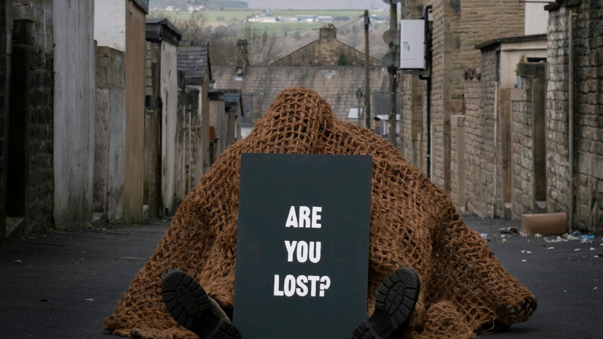 A figure sits in an alleyway of a Northern village, with fields and hills in the distance. They are covered in a rope net and holding up a chalkboard sign with the words ‘ARE YOU LOST?’ written on it.